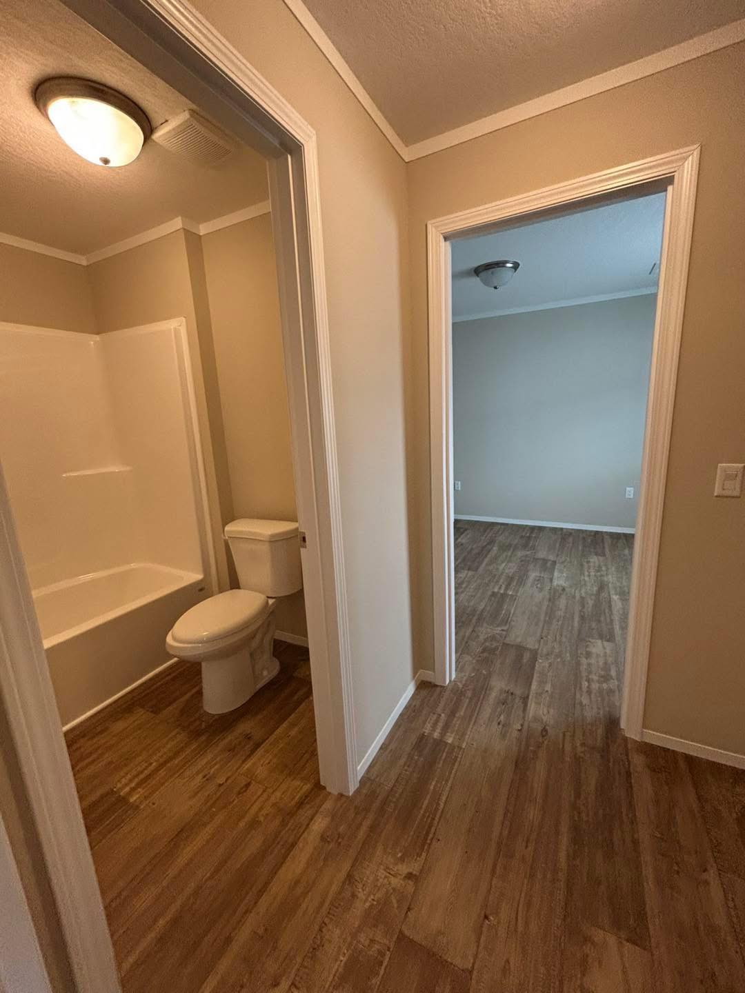 A hallway with wood floors leads to a bathroom on the left, featuring a bathtub and toilet, and an empty room with beige walls straight ahead. Bright, clean, and minimalistic.