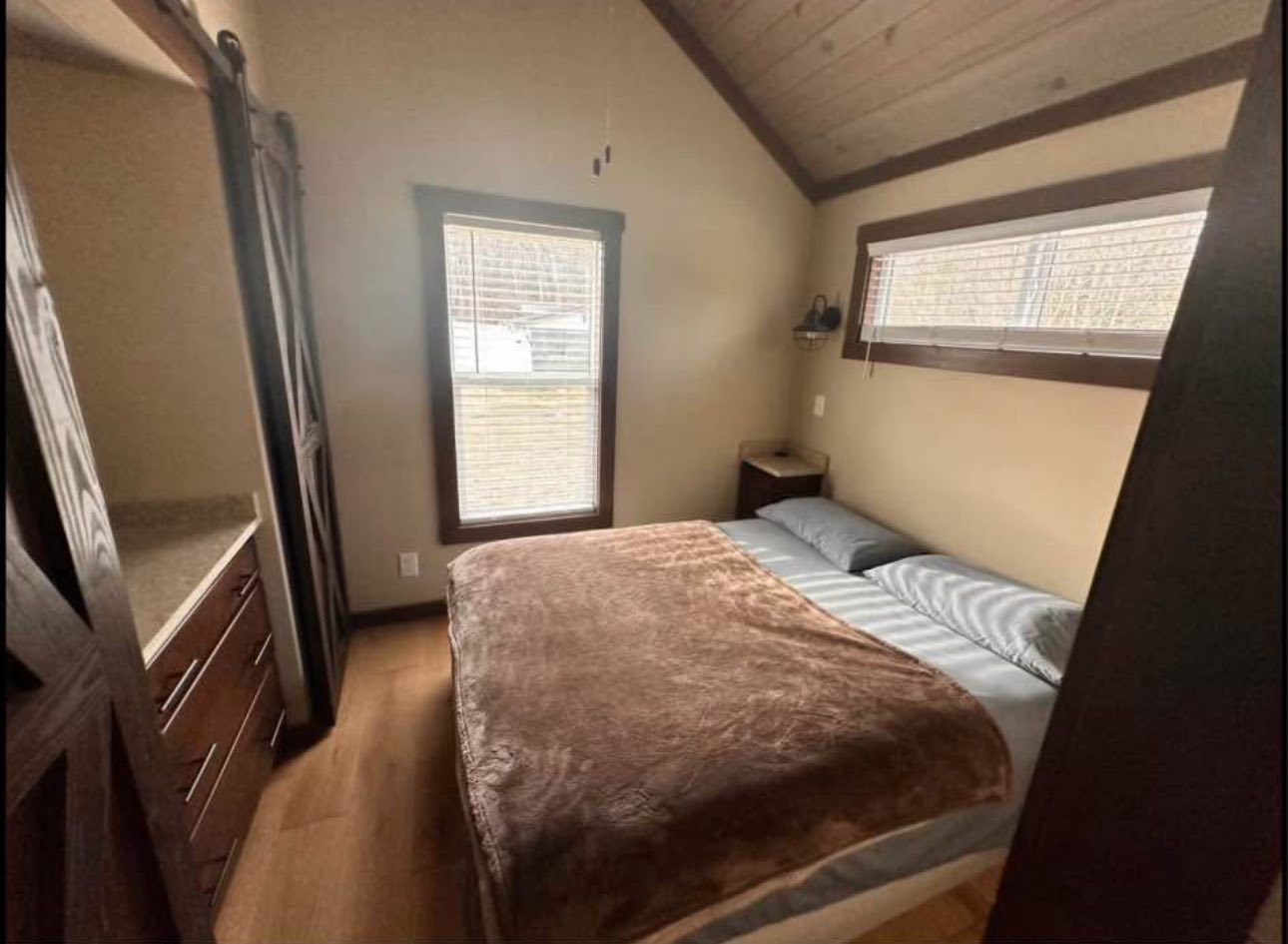 Cozy bedroom with sloped ceiling, featuring a single bed with a brown blanket, two small windows, and dark wooden cabinets on the left. Warm, inviting atmosphere.