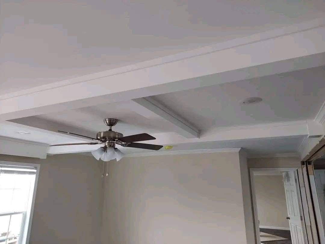 Ceiling fan in a beige room with a coffered ceiling design, near a window with natural light. The room feels modern and spacious.