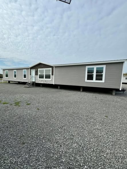 A large, gray mobile home with white trim sits on gravel against a cloudy sky. It features multiple windows and a small set of entrance steps.