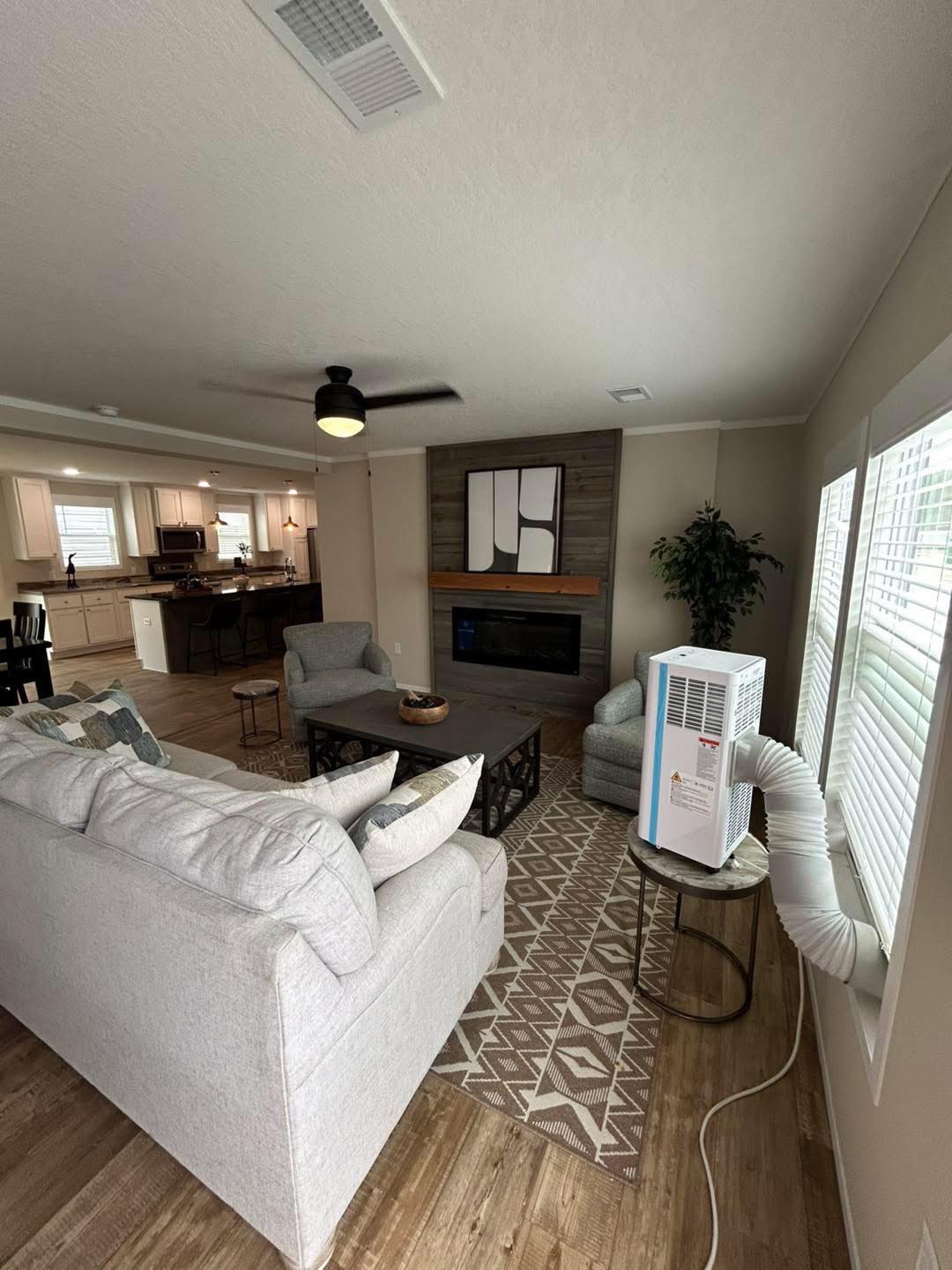 Modern living room with a gray sofa, armchairs, a geometric rug, and a coffee table. The room is bright with large windows and an air purifier. A kitchen with wooden cabinets is visible in the background. The atmosphere is cozy and elegant.