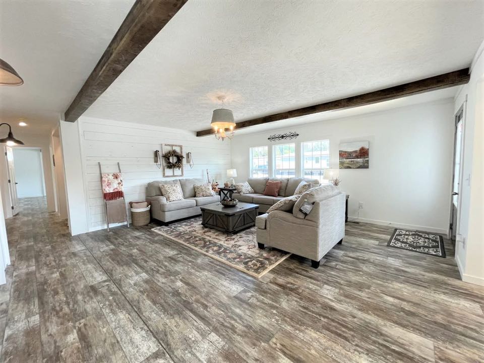 Modern farmhouse living room with rustic wood beams, gray sectional sofa, and cozy decor. Natural light from large windows on patterned rug.