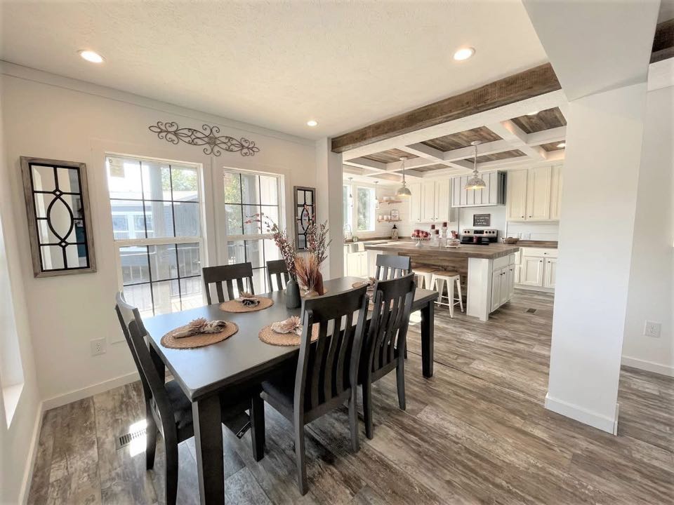 Bright dining area with a dark wooden table for six, next to large windows. Rustic kitchen in background features wood beams and modern appliances.