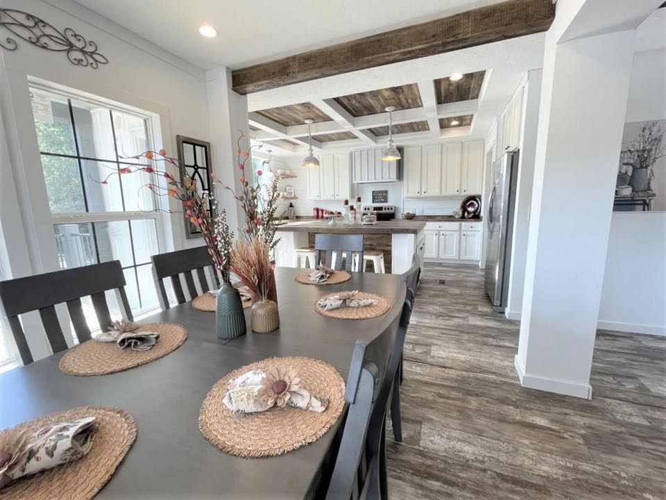Modern dining room and kitchen with a rustic vibe. A large table features woven placemats and napkins. Exposed beams and wooden flooring add warmth.