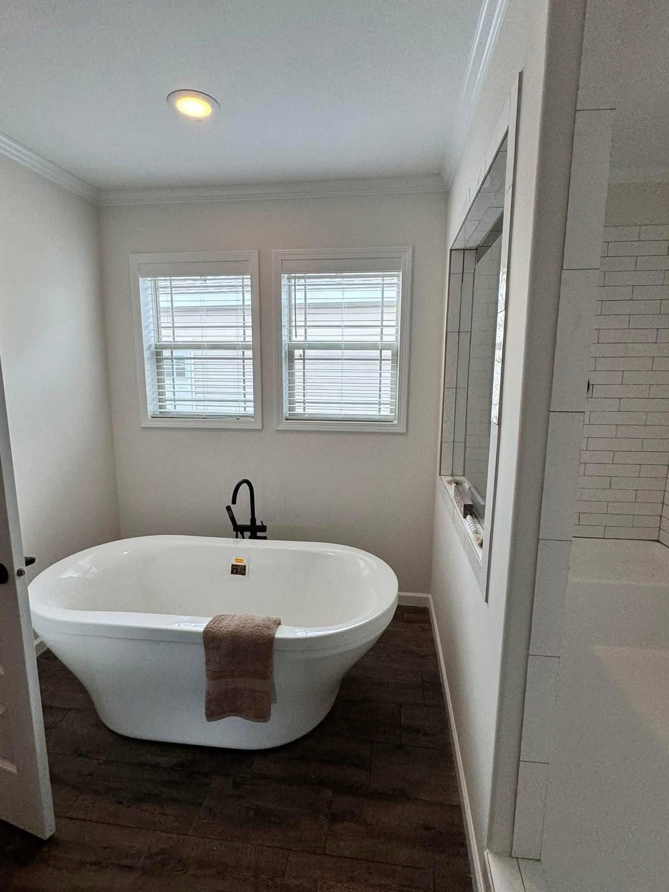 Elegant bathroom featuring a white freestanding tub with a dark faucet, set against white walls and two windows, on a dark wood floor.