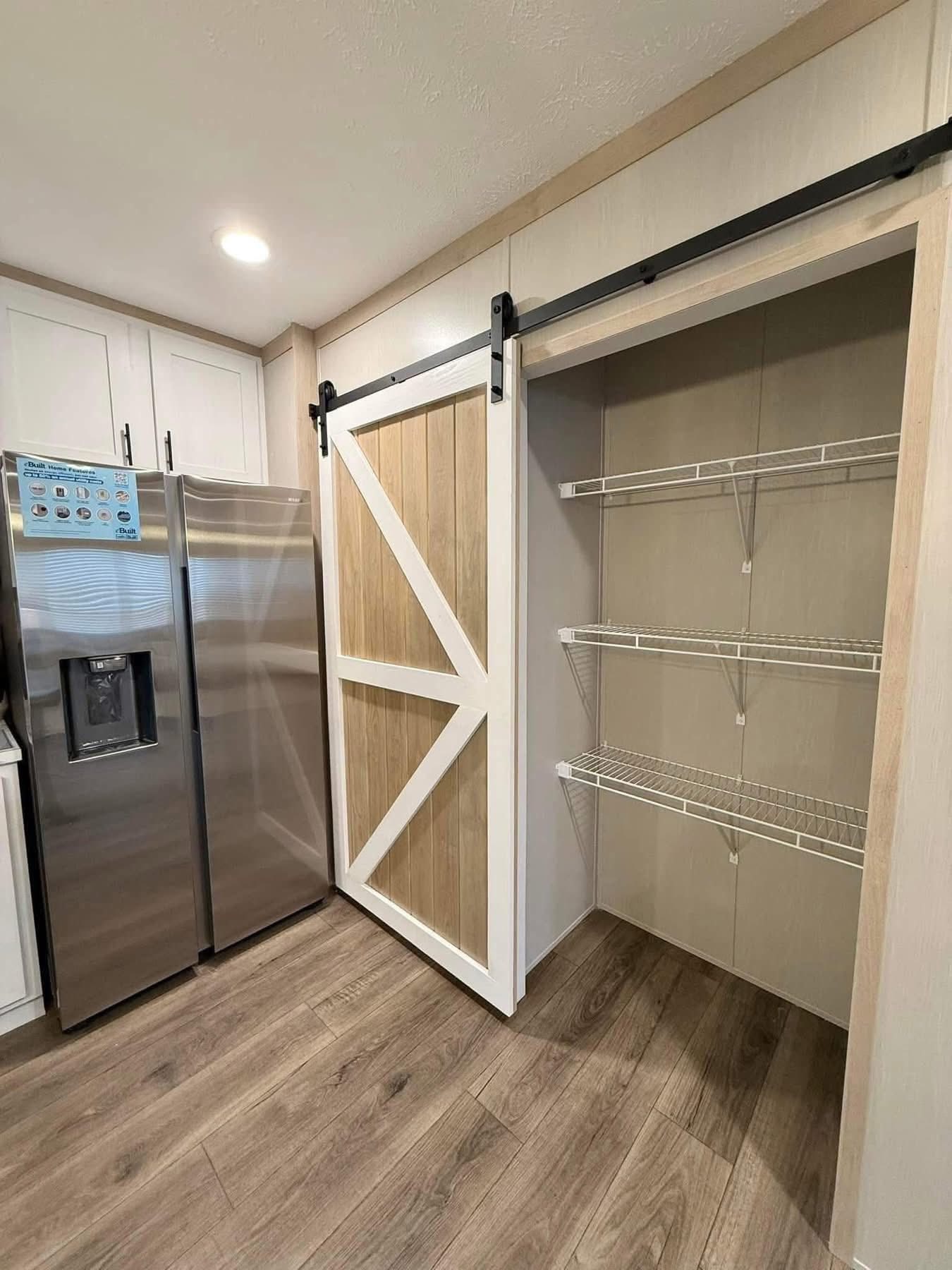 Modern kitchen with stainless steel fridge on the left, a sliding barn door with wood accents in the center, and open wire pantry shelves on the right. Warm ambiance.