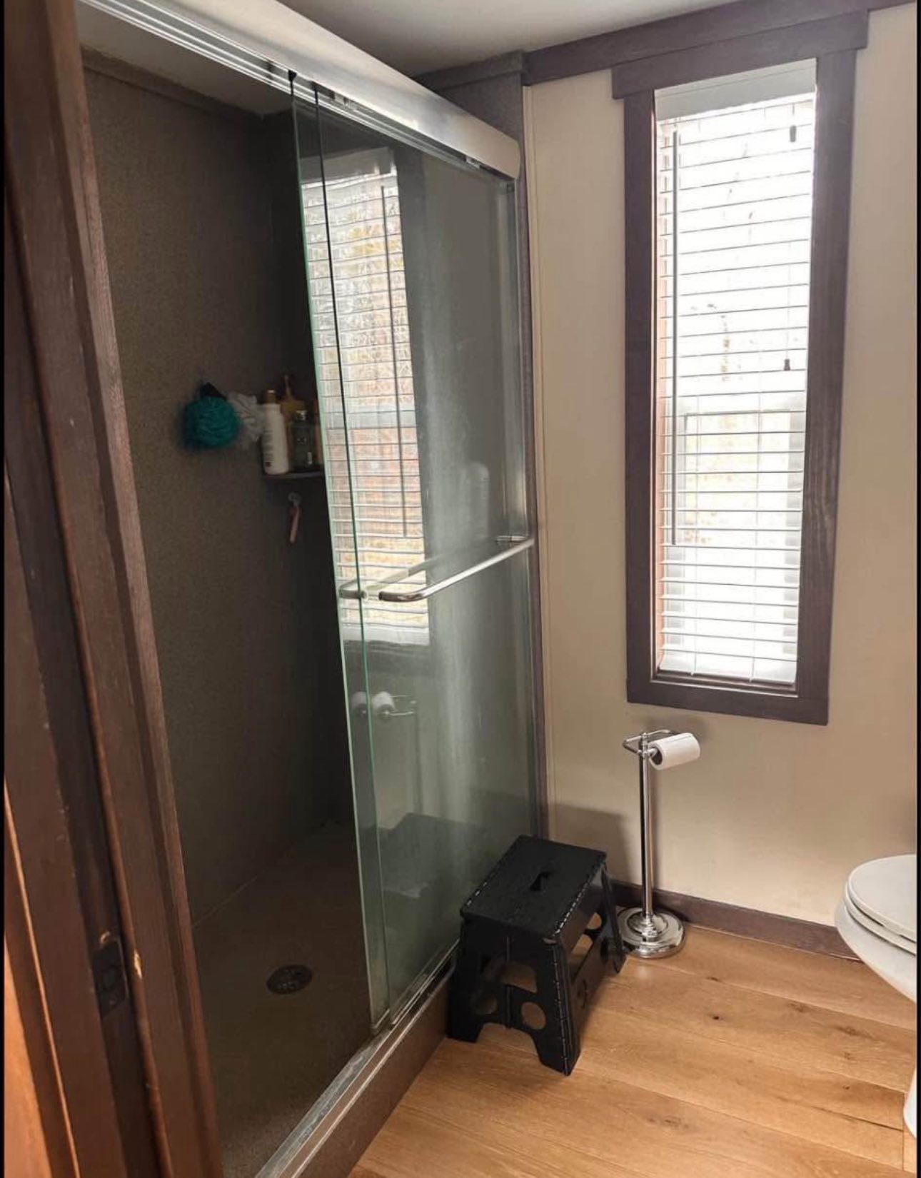 A bathroom with a glass-enclosed shower, wooden floors, and a window with blinds. There is a black stool and a roll of toilet paper on a stand nearby.