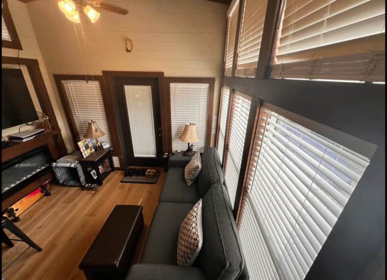 Cozy living room with a gray couch, patterned pillows, and a wooden coffee table. Large windows with blinds allow warm light, creating an inviting atmosphere.