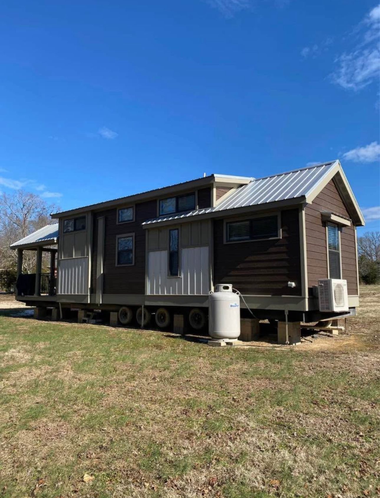 A tiny house with a metal roof sits on a grassy field under a clear blue sky. The house is elevated, with brown siding and large windows, creating a cozy vibe.