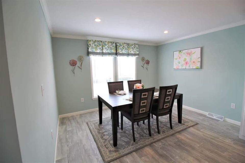 A serene dining room with pale green walls features a dark wooden table, four chairs, floral artwork, and patterned curtains over a large window.