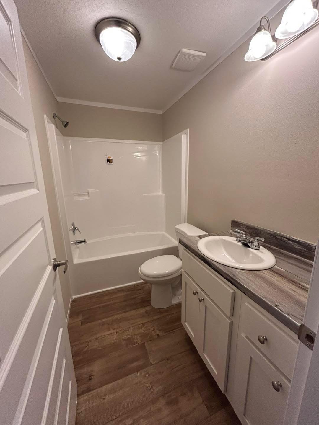 Compact bathroom with a white bathtub and shower, modern vanity with a sink, wooden cabinets, wood floor, and bright lighting. Minimalist and clean design.