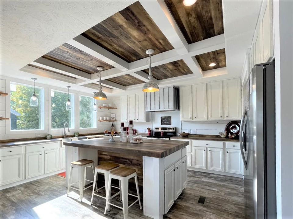 Spacious kitchen with rustic wooden ceiling panels, white cabinets, and a central island with stools. Large windows provide ample natural light.