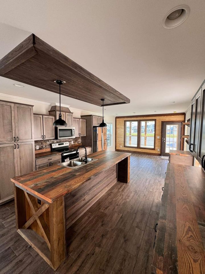 Rustic kitchen with wooden cabinetry and floors. Central island features a sink, pendant lights above. Sunlit doors lead to a porch outside. Cozy ambiance.