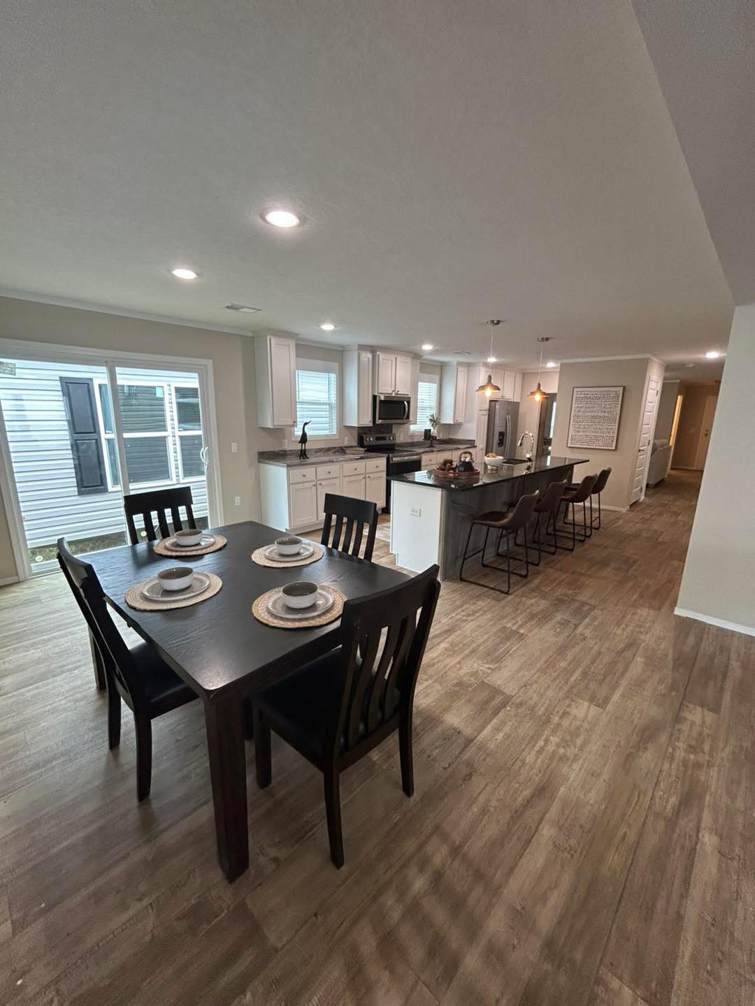 Spacious kitchen and dining area with wood flooring. A dark dining table set for four is in the foreground, with a modern kitchen island and barstools behind.