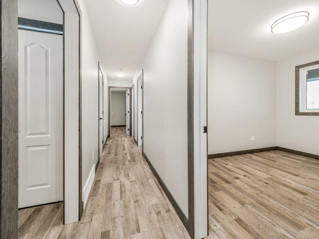 A bright, modern hallway with light wood flooring and white walls, leading to multiple doors. A room to the right adds depth and openness.