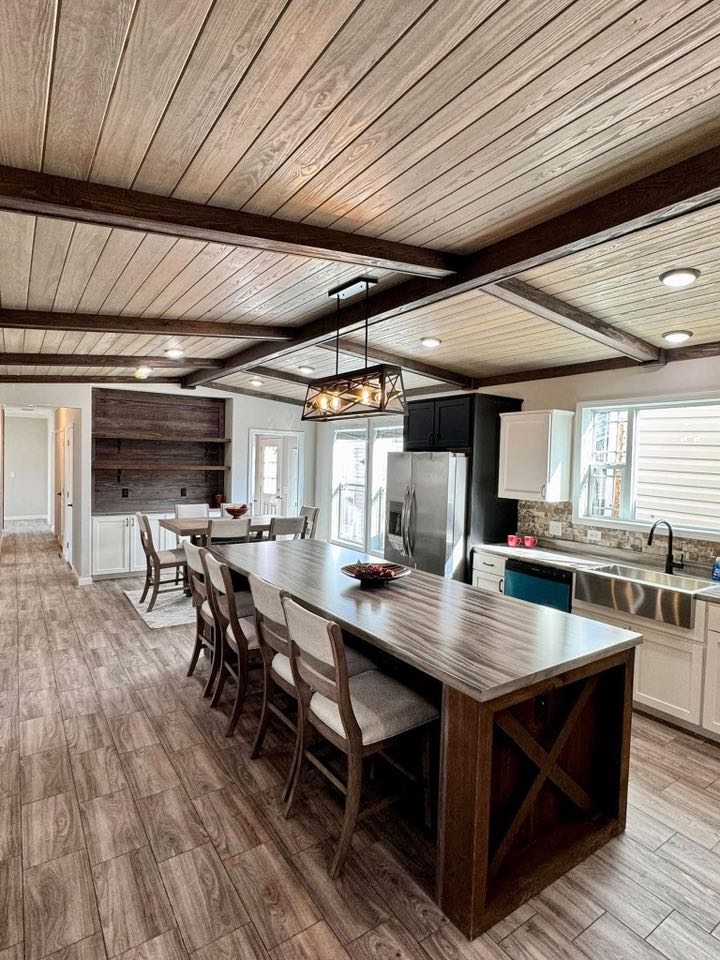 Spacious kitchen with wooden ceiling beams and floor, a long island with chairs, stainless steel appliances, and bright natural lighting. Cozy ambiance.
