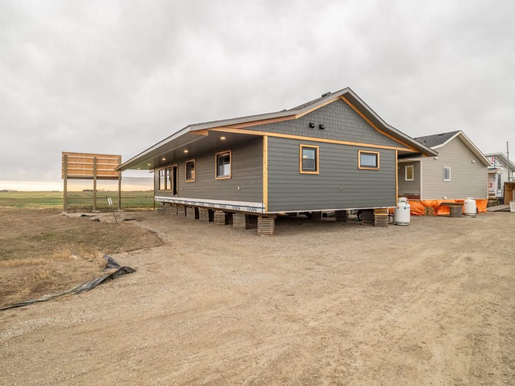 Elevated gray house under construction on a gravel lot, surrounded by open fields. Overcast sky creates a subdued and calm atmosphere.