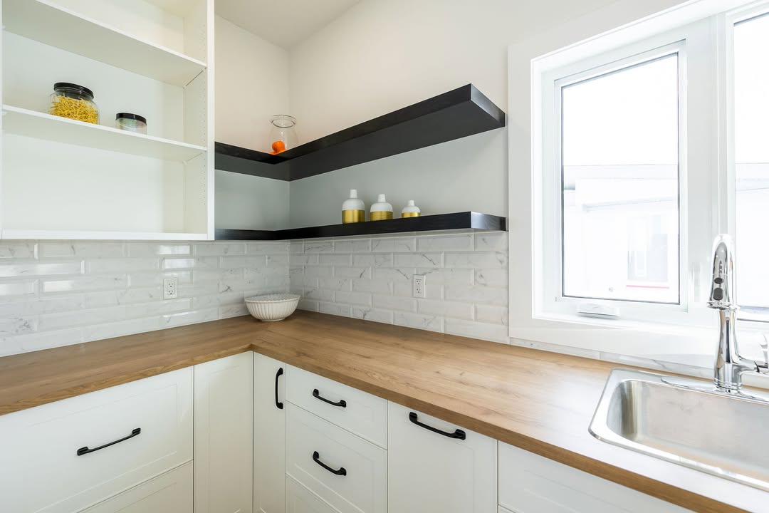 Modern kitchen with white cabinets, wooden countertops, and a stainless steel sink. Black shelves hold containers; a bright window adds natural light.
