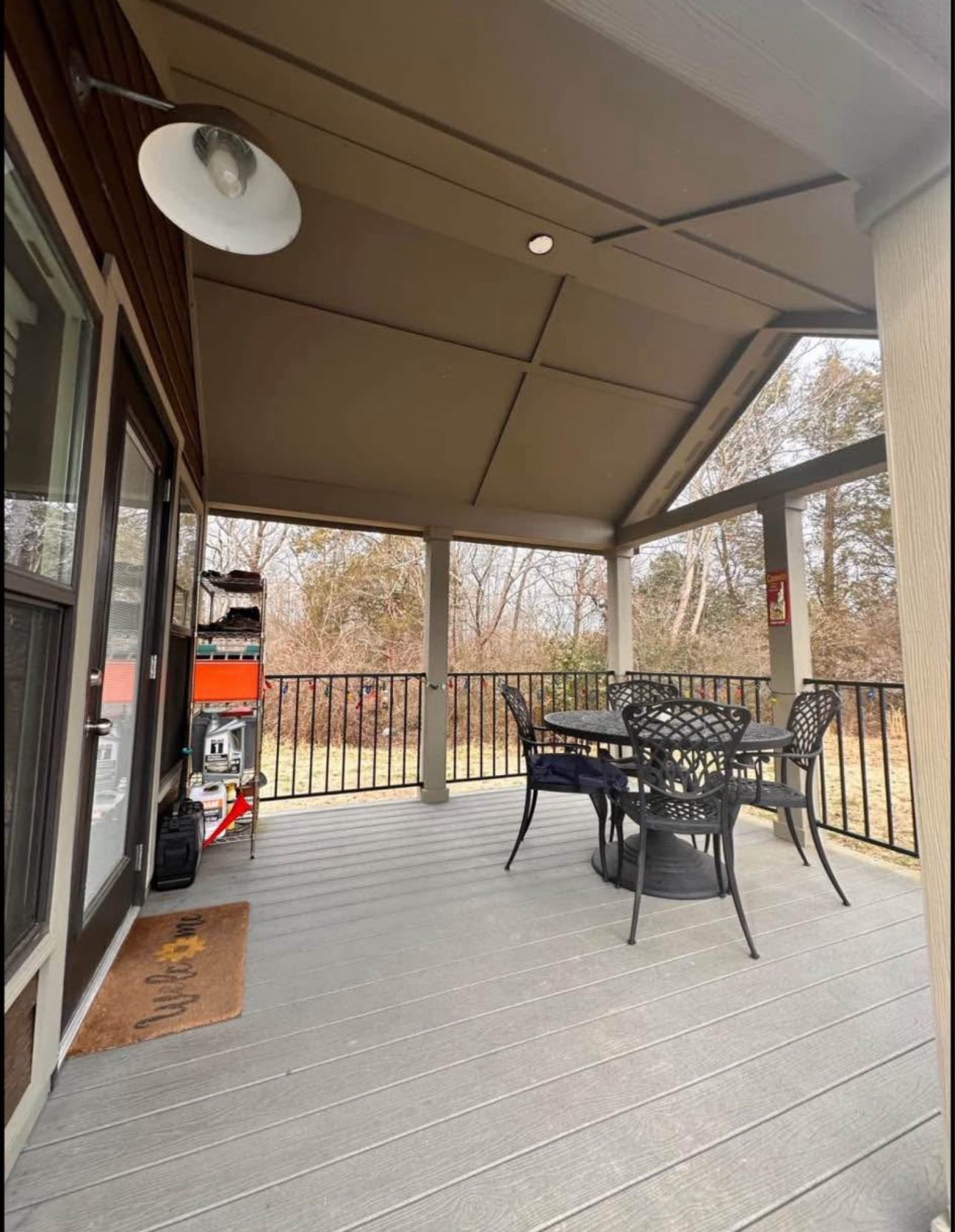 Covered patio with a black metal table and four chairs on a gray deck. Surrounded by trees, it conveys a calm, cozy outdoor setting.
