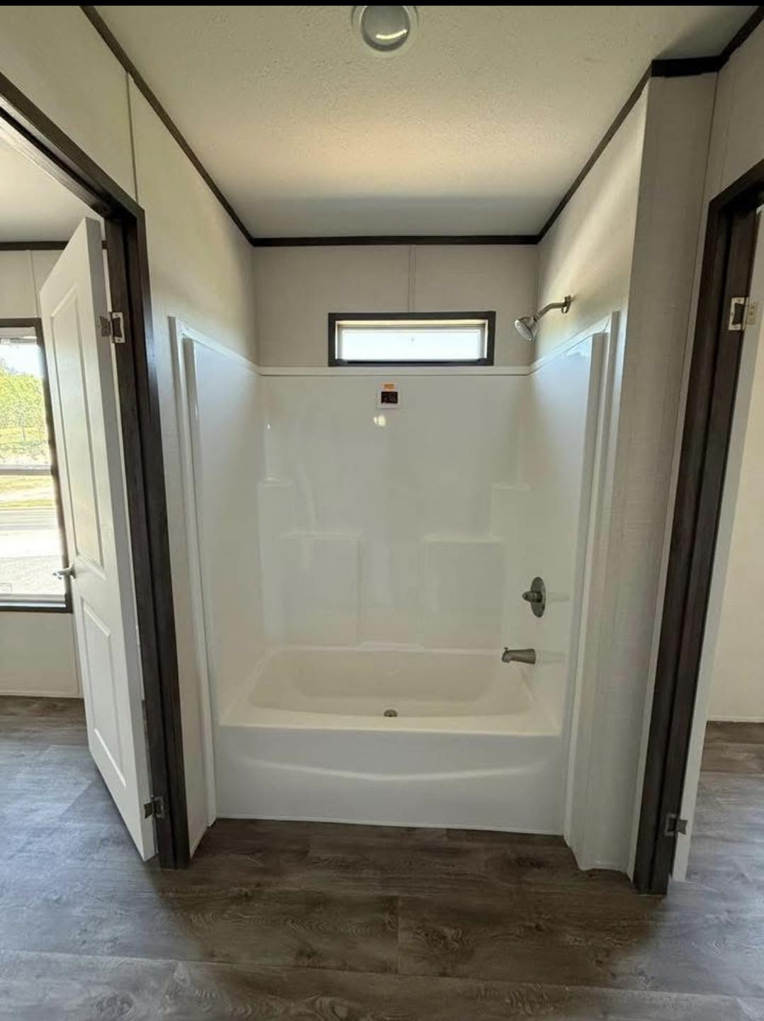 Compact bathroom with a white bathtub, surrounded by light walls and a small window above. Dark wood doors open to reveal the space, adding contrast.