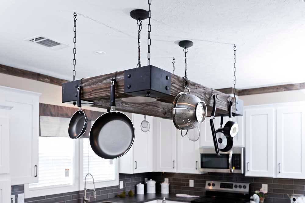 Hanging wooden pot rack with black metal chains holds various kitchen utensils, like pans and colanders. Modern kitchen with white cabinets in the background.
