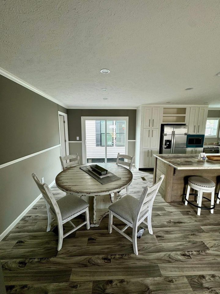Cozy dining area with a rustic round table and four white chairs on wooden flooring, adjacent to a modern kitchen with white cabinets and island. Soft natural light.