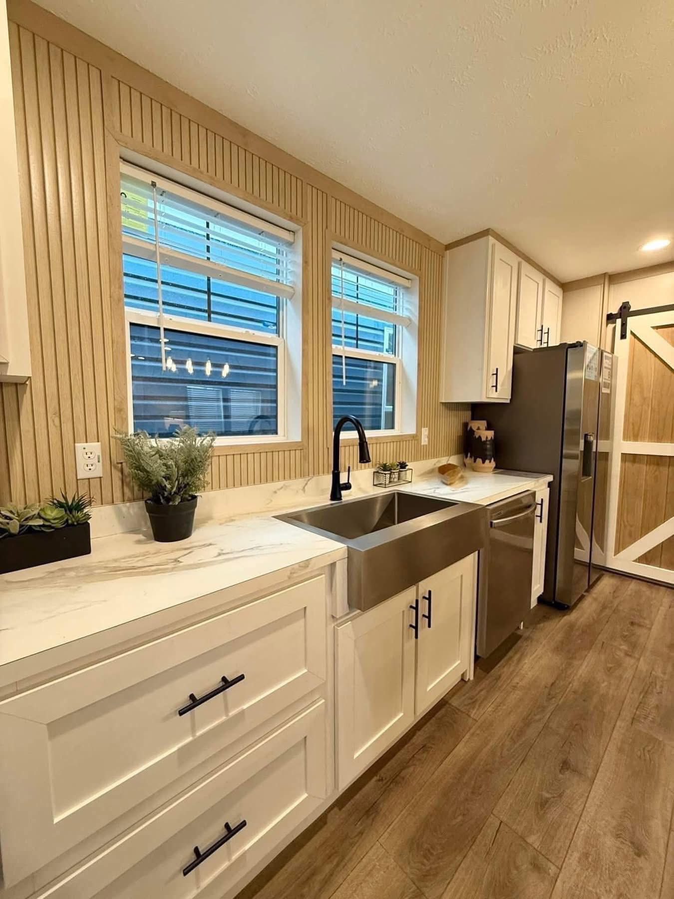 Modern kitchen with a farmhouse vibe: white cabinets, stainless steel sink and appliances, light wood floors, barn-style door, and bright windows.