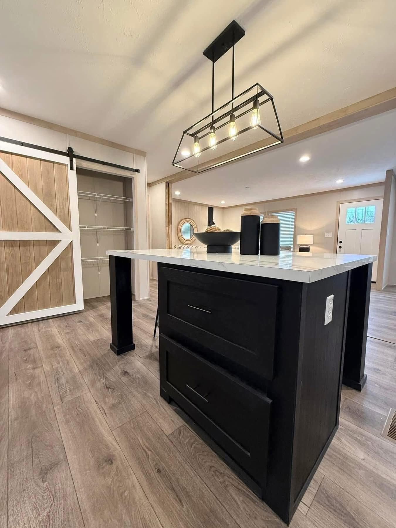 Modern kitchen with a black island and white countertop, pendant light, and open pantry with a sliding barn door. Warm wood floors enhance the contemporary feel.