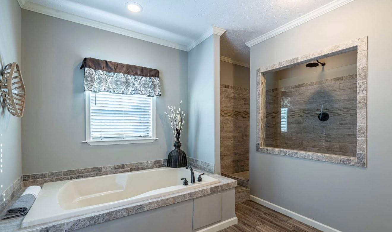A modern bathroom features a white bathtub with tile accents, a potted plant, and a decorative valance above a window. There's a spacious shower with a glass partition. The atmosphere is clean and tranquil.