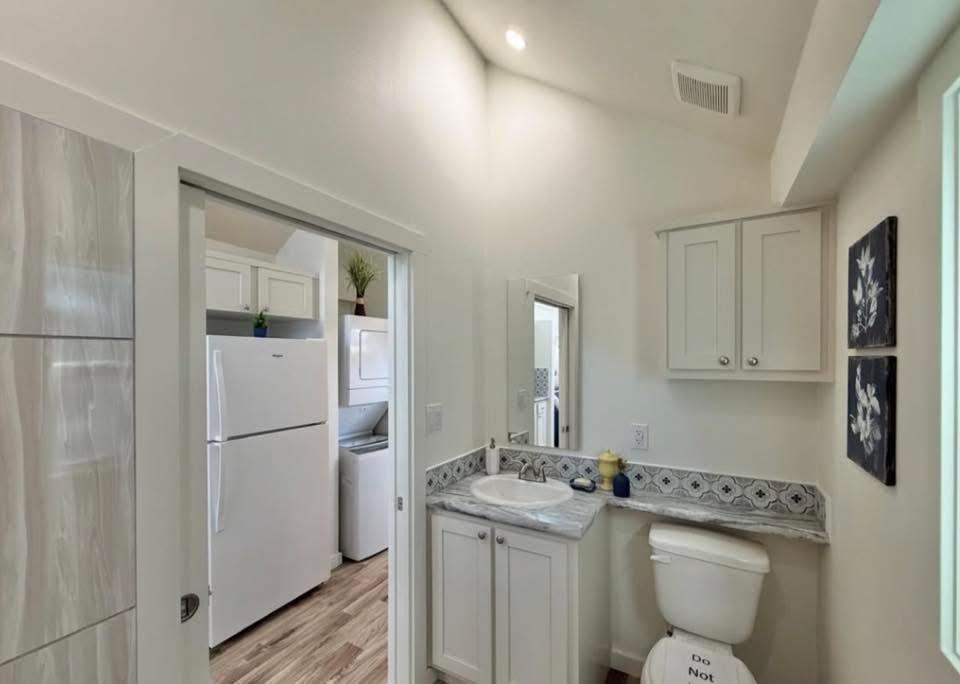 A small, bright bathroom with white cabinets and a marble countertop. Art hangs on the wall above the toilet. The adjacent laundry room is visible.