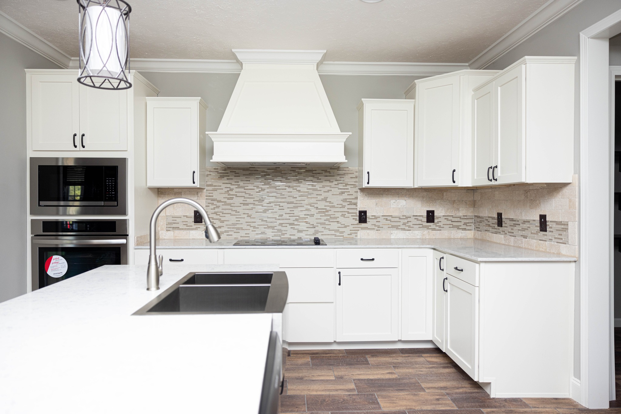 Bright modern kitchen with white cabinets, stainless steel appliances, and a stone backsplash. A large sink and an elegant pendant light enhance the clean, sophisticated look.