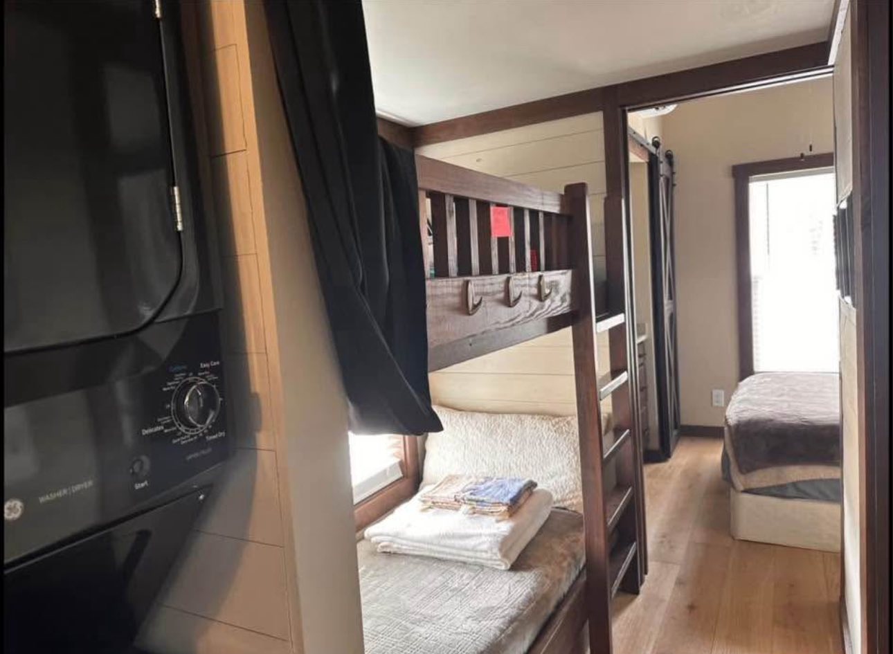 Compact interior with wooden bunk beds on the right, a cozy bed with folded towels beneath. Laundry area on the left, bright natural lighting. Warm and inviting.
