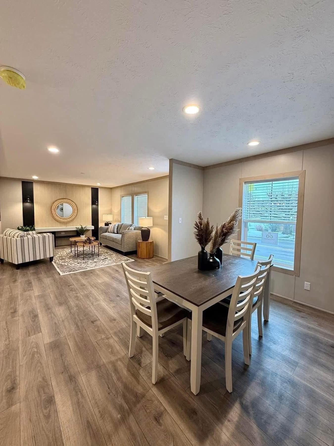 Modern living and dining area with wooden floors. Light-colored walls, table with chairs, pampas grass in vase. Cozy seating, large window, warm lighting.