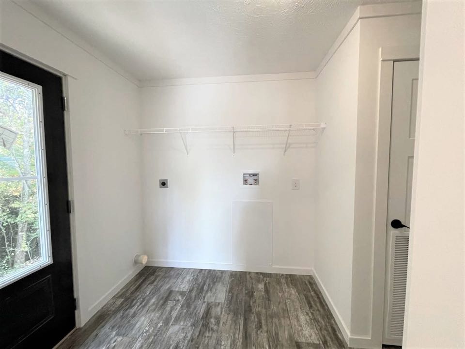 A bright, empty laundry room with white walls and a gray wood floor. It features a wire shelf, connection points for appliances, and a glass-paned door.