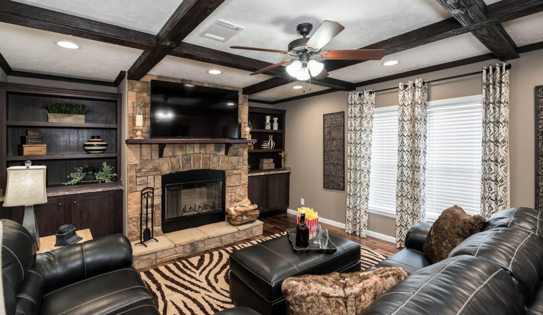 A cozy living room features a stone fireplace with a TV above, flanked by dark wood shelves. Black leather sofas face a zebra-patterned rug, creating a warm, inviting ambiance.