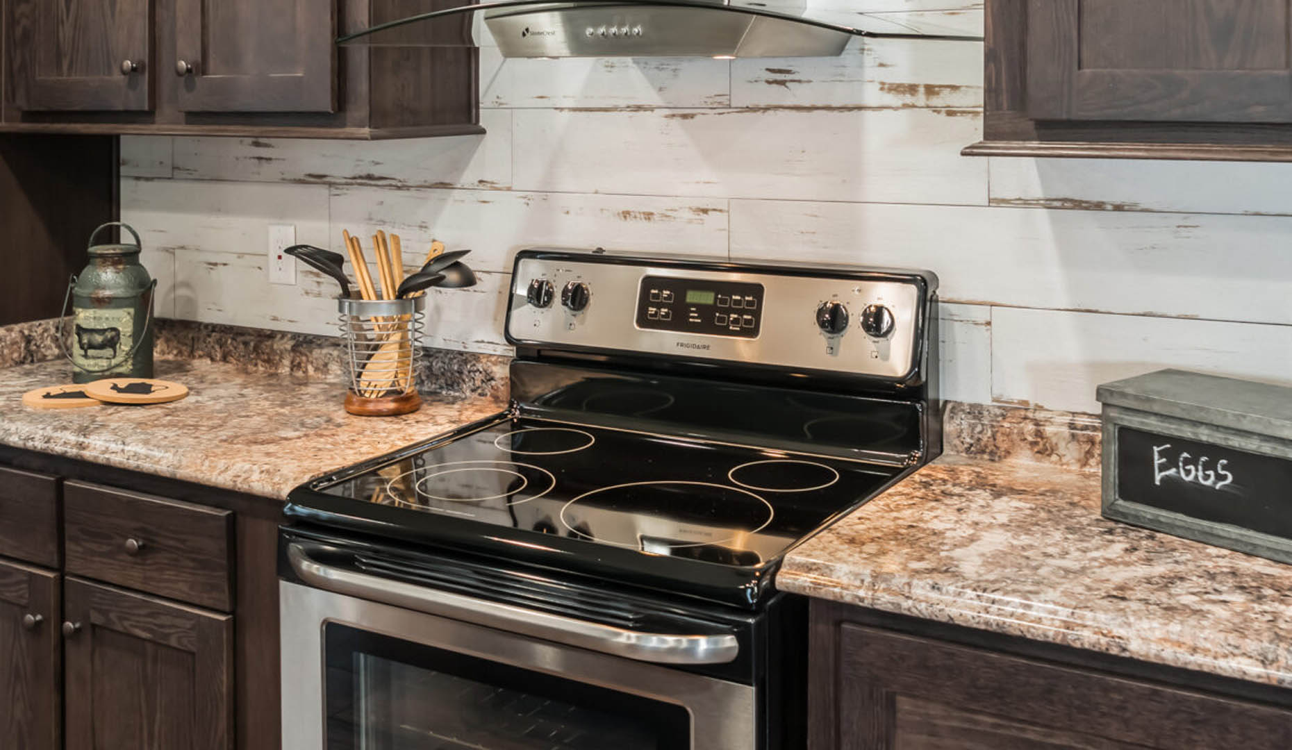A modern kitchen with dark wood cabinets and speckled countertops. A stainless steel stove is centered, surrounded by rustic decor and utensils.