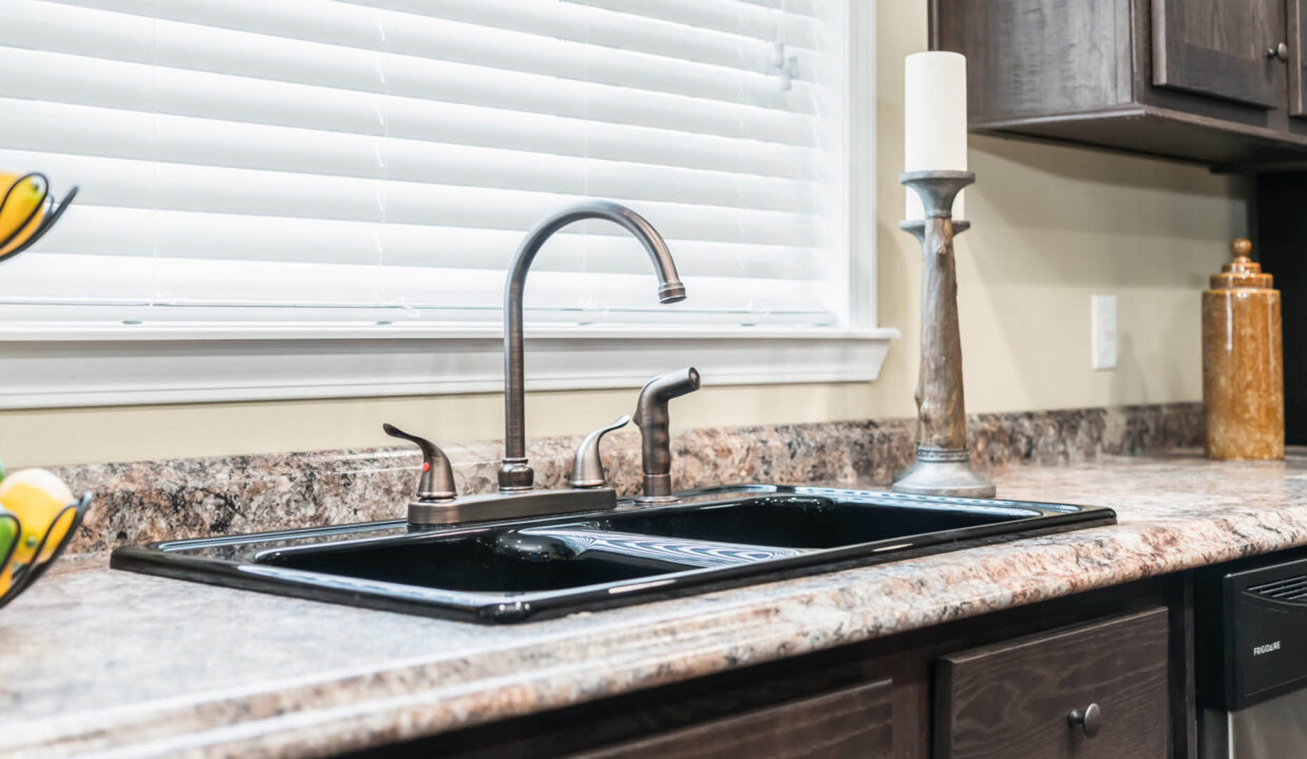 Modern kitchen sink with chrome faucet on marble countertop, flanked by a tall candle holder and decorative jar. Soft, natural light enters through blinds.