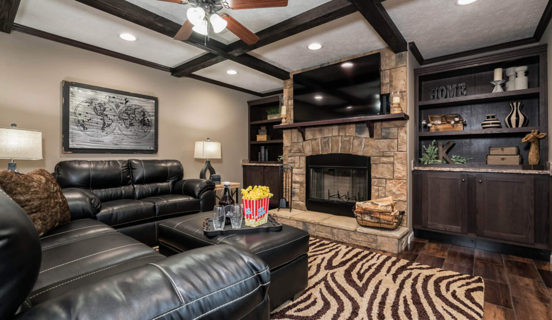 Cozy living room with black leather sofas, zebra-patterned rug, stone fireplace, and wooden shelves. A tray with popcorn and drinks on the ottoman.