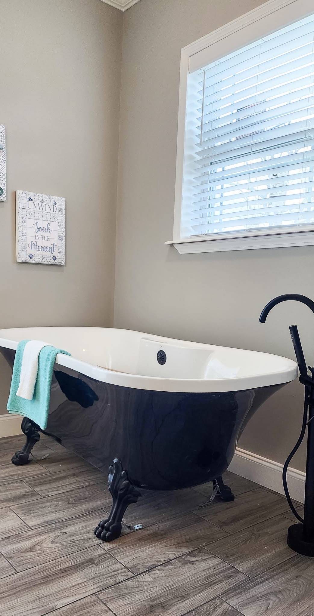 A vintage black clawfoot bathtub with a white interior and teal towel is set against beige walls and wooden flooring. A window with blinds and wall art are visible.