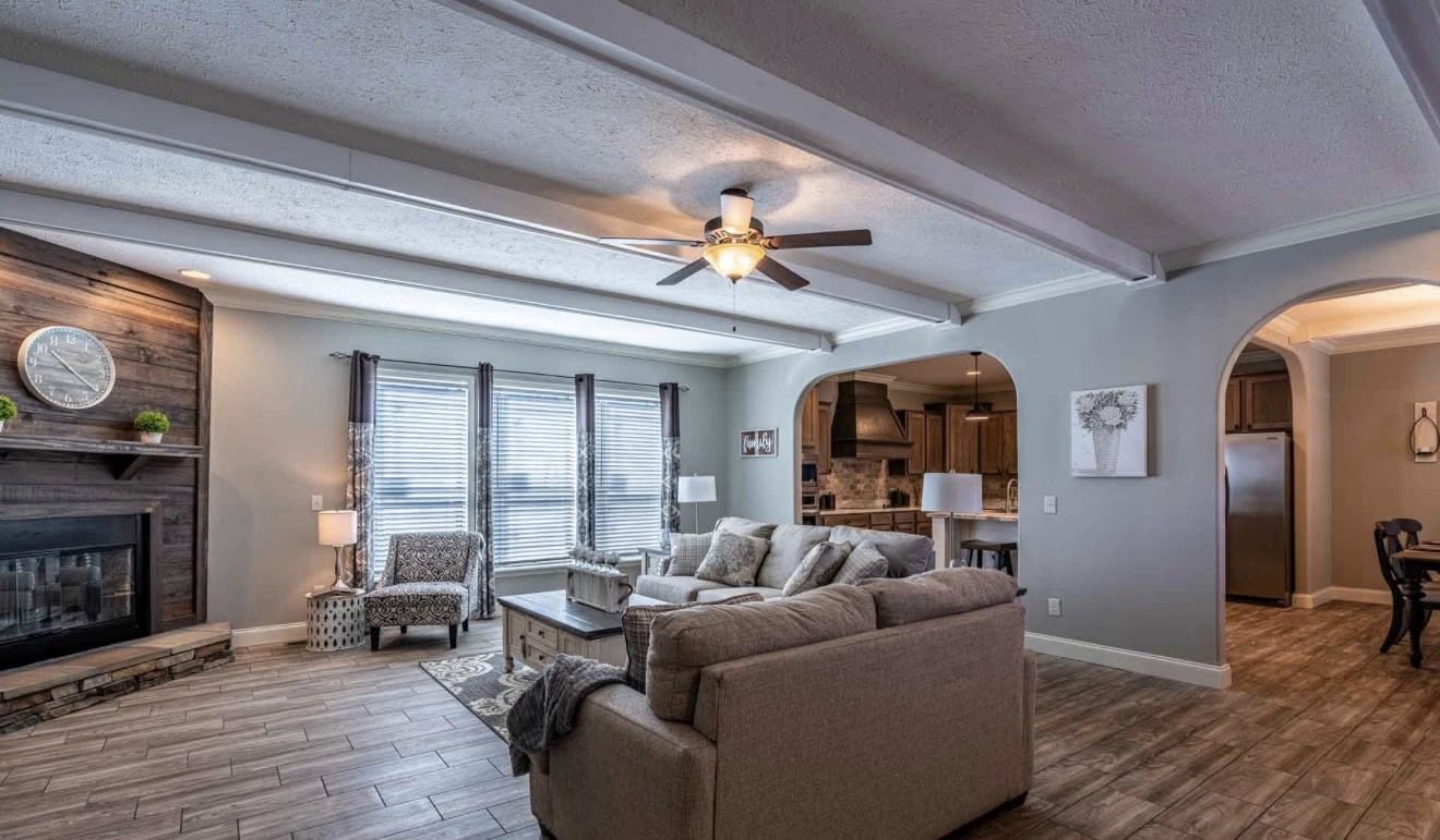 Spacious living room with light gray walls and a wooden floor. Features a beige sectional sofa, a stone fireplace, large windows, and ceiling fan. Cozy and inviting.