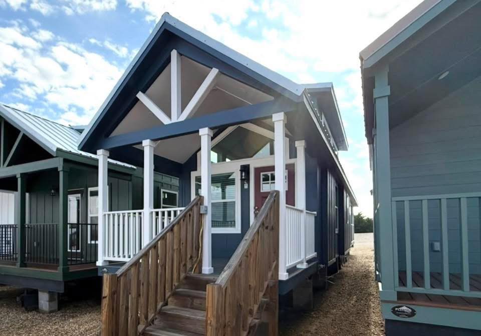 A small blue house with a gabled roof and white trim stands under a partly cloudy sky. Wooden steps lead to a porch with white railings, creating a cozy feel.