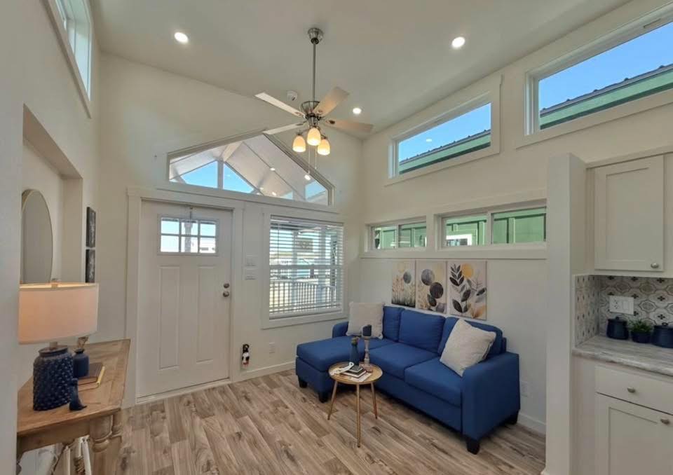 Bright, modern living room with high ceilings featuring a blue sofa, decorative pillows, and wall art. Large windows let in ample natural light. Boutique corner reveals cozy decor.