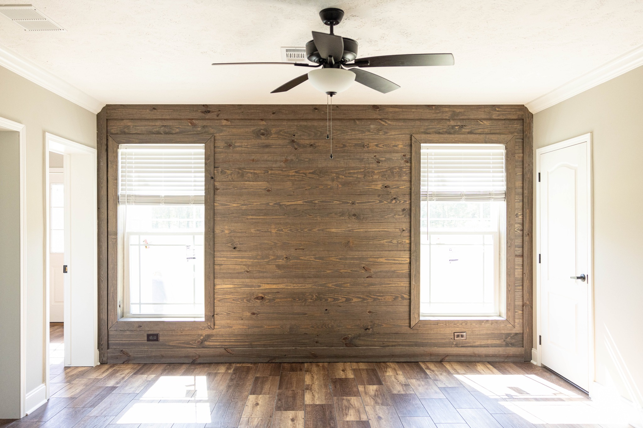 An empty room features a rustic wooden accent wall with two windows, a ceiling fan, and wooden floors. Sunlight streams, creating a bright, inviting space.