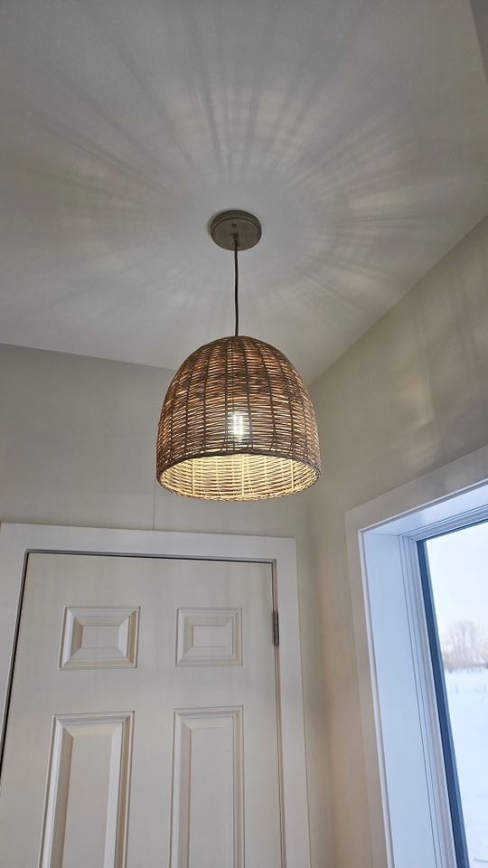 A woven wicker pendant light hangs from a beige ceiling, casting intricate shadows. It is near a white door and a window with a winter scene outside.
