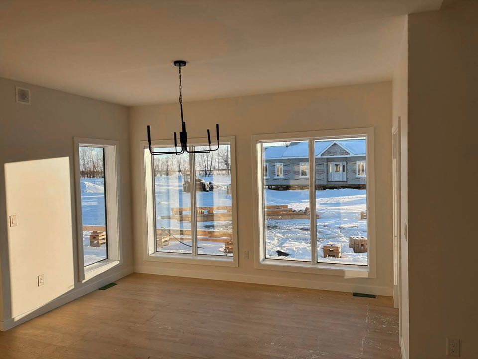 Sunny room with light wood floor and large windows overlooking a snowy landscape and houses. A modern black chandelier hangs from the ceiling.