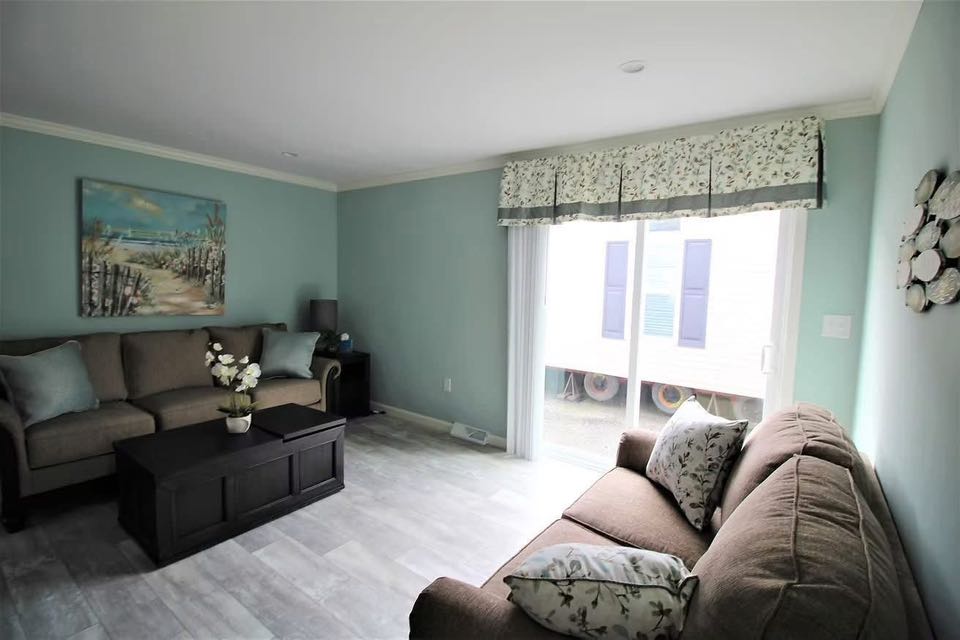 A cozy living room with light blue walls and soft gray flooring. Two taupe sofas adorned with patterned pillows face a central dark wood coffee table. Decorative art hangs behind one sofa, and sheer curtains frame a large window, flooding the room with natural light. The atmosphere is calm and inviting.