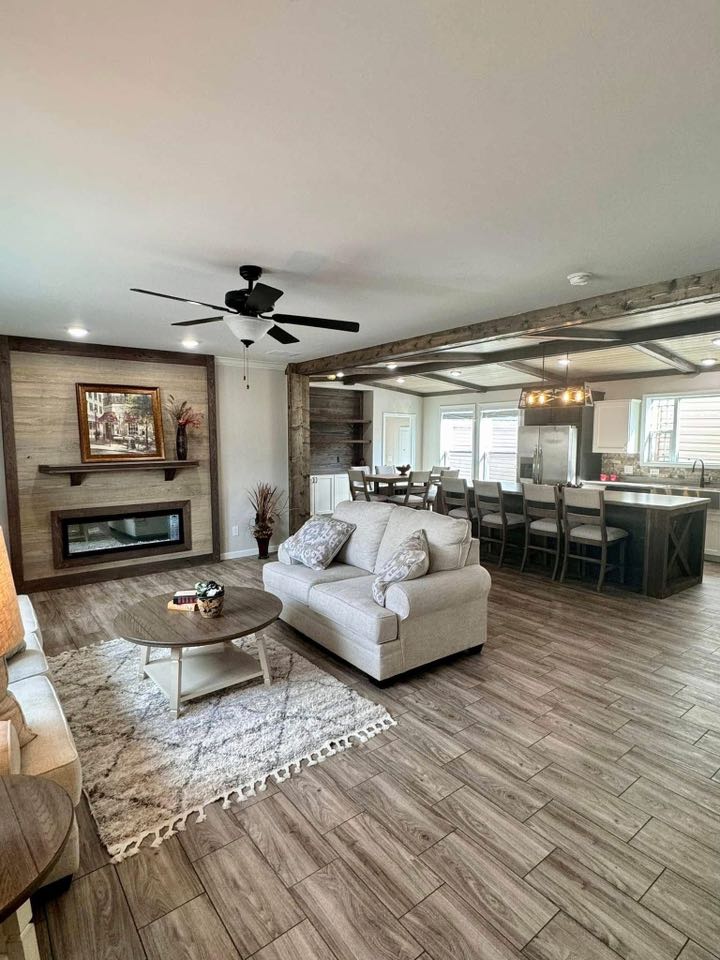 Spacious living room with light wood flooring, beige sofa, and coffee table on a patterned rug. An open kitchen with barstools and modern decor nearby.