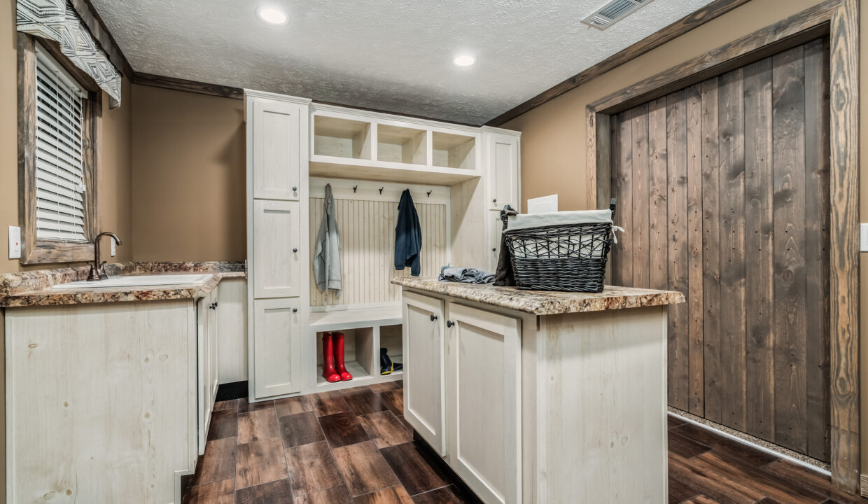 A cozy mudroom with beige walls and a wooden floor features a laundry sink, cabinets, and a bench with hooks. A basket sits on a central island.