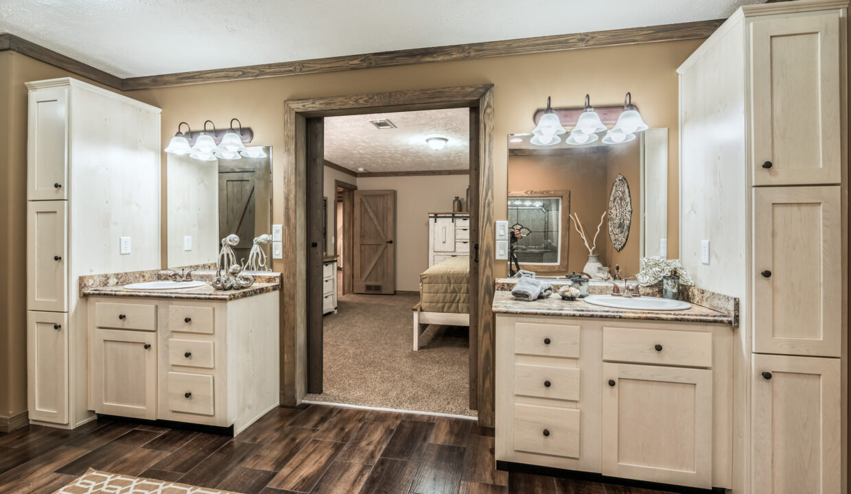 Rustic bathroom with dual sinks, stone countertops, and wooden cabinets. Warm lighting and wooden floor. Bedroom visible through the doorway. Cozy atmosphere.