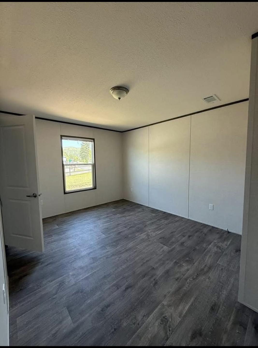 Empty room with light gray walls, dark wood flooring, and a small window showing a sunny outdoor view. An open door on the left suggests entry.